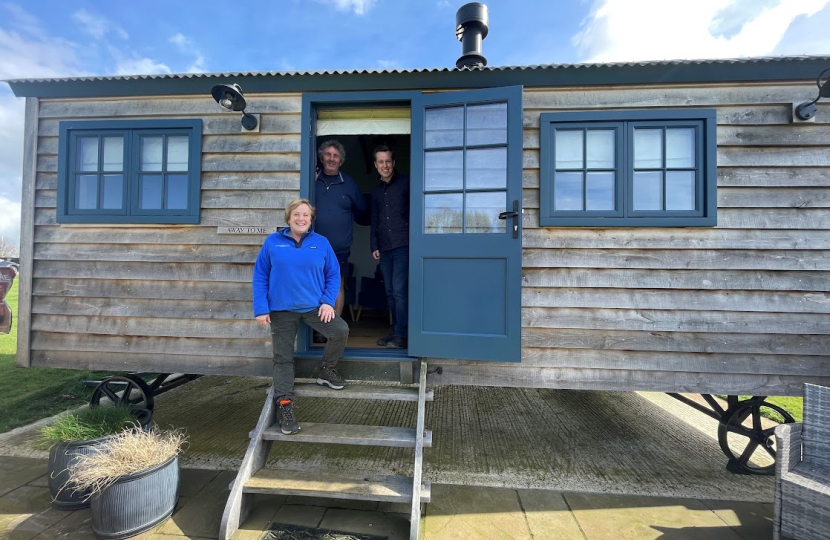 Tom with two representatives at New Lodge Farm in Bulwick peering out of the front door of a wooden static property. All three are smiling as they look out.
