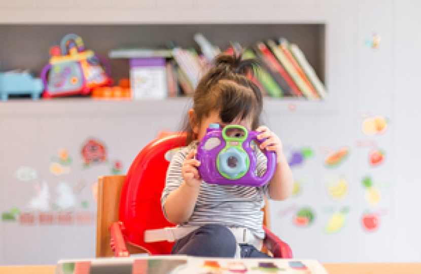 A young girl sat in a high chair. She is looking through a toy camera. In the background, there are colourful stickers on the wall and lots of books and toys on the shelf.