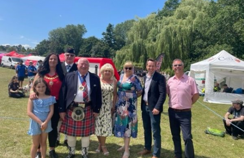 Tom stood with a number of attendees of the Highland Gathering in Corby, including the Mayor of Corby and NNC councillors. There are stalls in the background and a bright blue sky.