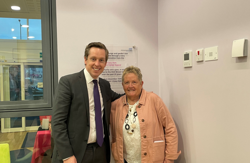 Tom Pursglove MP and Glennis Hooper, who spearheaded the 'Crazy Hats' project at Kettering General Hospital, in front of the plaque marking the opening of the new waiting lounge. Both are smiling while Tom puts his arm around Glennis' shoulder.