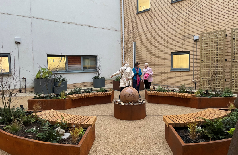 A group of people looking around the new 'Crazy Hats' Garden at Kettering General Hospital. A round stone fountain centrepiece reads 'Hope.' Wooden benches surround it with freshly planted shrubs behind them.