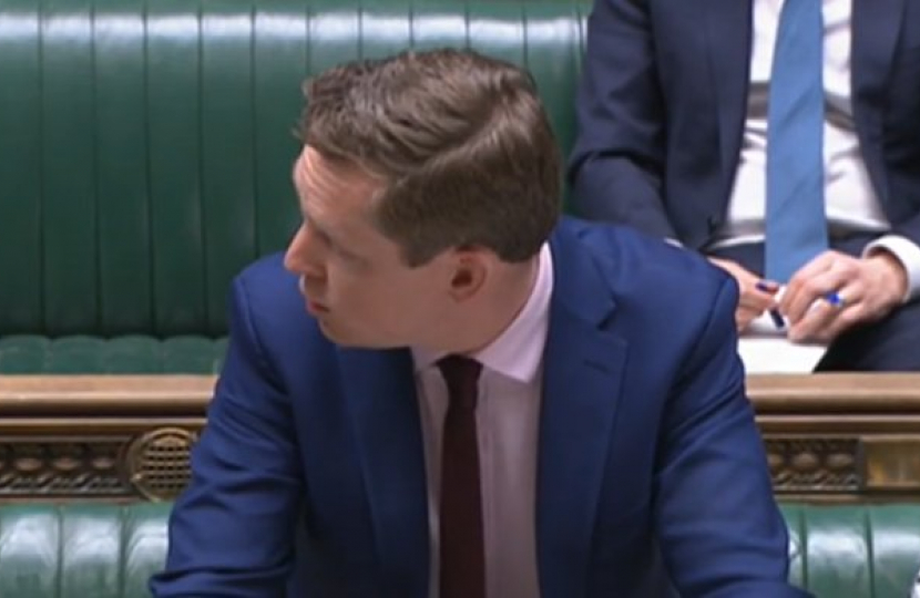 Tom Pursglove MP, in his role as Minister for Disabled People, Health and Work, is stood at the Dispatch Box in the House of Commons. He is looking over to his right, answering a question from a colleague. Other colleagues sit around him on the green benches.