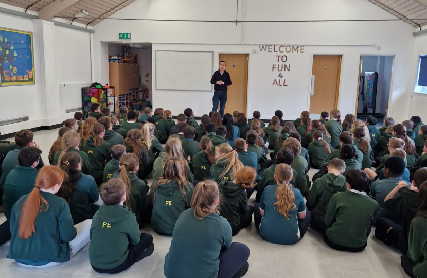 Tom stood in an assembly hall in front of Year 6 students, who are all sat down. The children are wearing a dark green uniform
