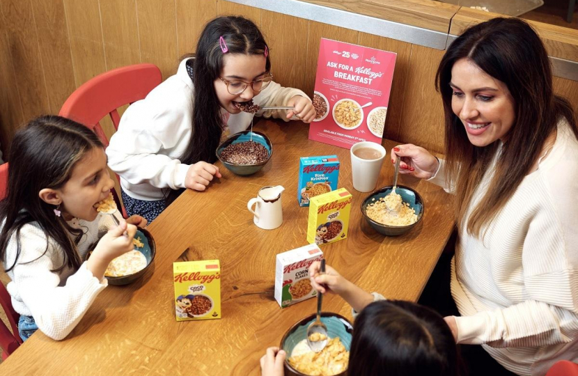 Three young children are sat at a table with their mother eating bowls of cereal