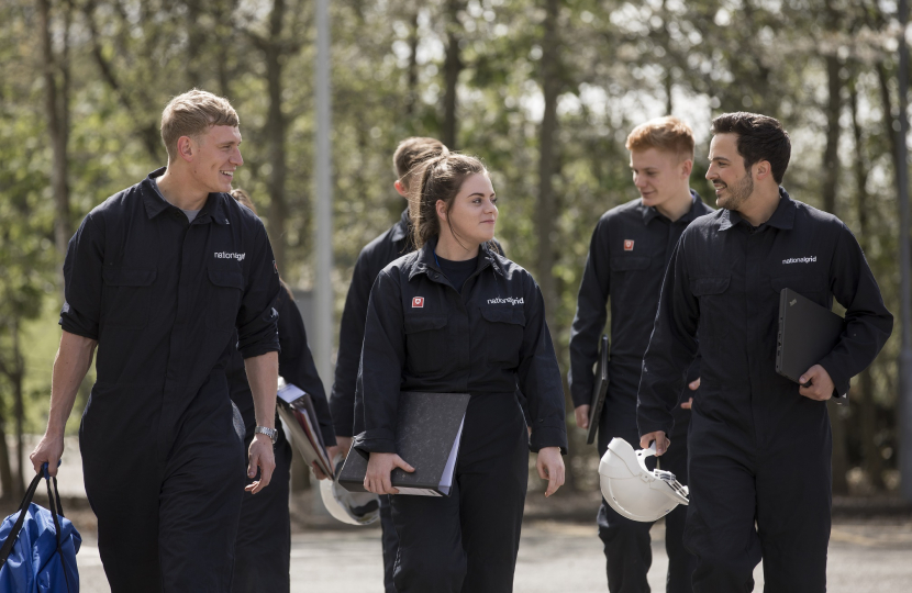 A group of young people participating in the National Grid Apprenticeship Scheme. They are each wearing black uniforms including the 'National Grid Logo' and some are carrying paperwork or a hard hat. They are smiling as they walk along, talking to one another.