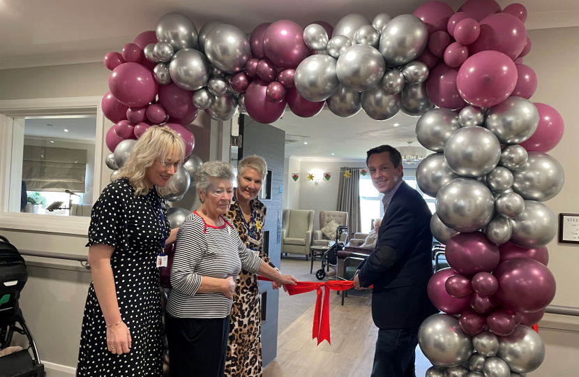 Tom Pursglove MP, with a group of residents and staff at Priors Hall Care Home about to cut the ribbon to officially open the new 'Steeltown' floor. The group are stood smiling beneath a bright pink and silver balloon arch.