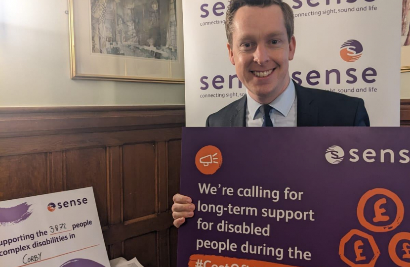 Tom holding a purple and orange poster by Sense which reads 'We are calling for long-term support for disabled people during the cost of living crisis. The is a banner behind Tom which the Sense logo on it.