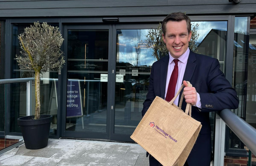 Tom standing in front of the care home holding a bag with the home's logo on it. He is smiling and wearing a suit with a red tie