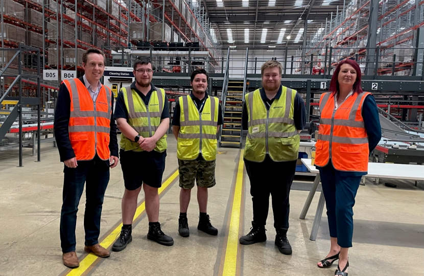 Tom stood with four employees of Wincanton. All are wearing hi-viz jackets and there is extensive warehouse racking in the background.