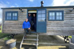 Tom with two representatives at New Lodge Farm in Bulwick peering out of the front door of a wooden static property. All three are smiling as they look out.