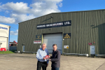 Tom and Andrew Howard, Managing Director of PC Howard, oustide a large, green warehouse. Above the door, it reads 'Howard Warehousing LTD.'