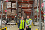 Tom and a member of staff from the Primark distribution facility in Islip, posing for a photograph. Behind them, boxes of stock are stacked up on pallets.