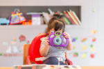 A young girl sat in a high chair. She is looking through a toy camera. In the background, there are colourful stickers on the wall and lots of books and toys on the shelf.