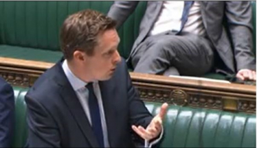 Tom stood in the House of Commons, answering a question. He gestures away from himself while doing so. 