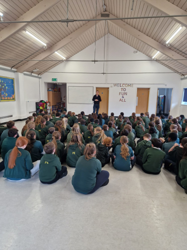 Tom stood in an assembly hall in front of Year 6 students, who are all sat down. The children are wearing a dark green uniform