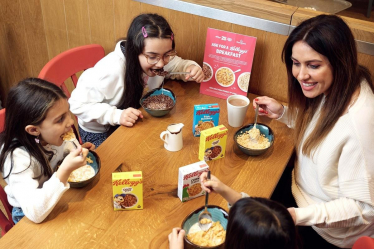 Three young children are sat at a table with their mother eating bowls of cereal