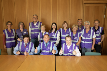 Tom Pursglove MP sat next to Philip Hollobone MP and Andrew Lewer MP giving a 'thumbs up' and wearing purple hi-viz jackets. Behind them are 10 individuals who volunteer as part of Northamptonshire Litter Wombles. Again, all are wearing purple hi-viz jackets. 