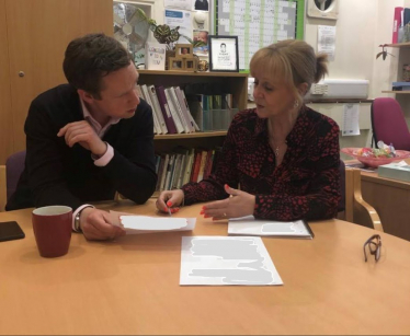 Tom Pursglove MP in a meeting with a representative from Pen Green. They are sat next to eachother reviewing some paperwork. The representative gestures to Tom as she is speaking. A mug and a black pair of glasses also sit on the table. A bookshelf features behind them.
