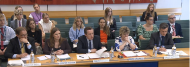 Image of Tom Pursglove MP sat next to other officials at the Select Committee meeting. They are sat at a long desk, with various papers in front of them. There are other individuals seated in the background watching the proceedings.
