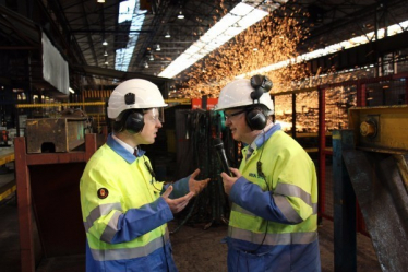 Tom Pursglove MP and a representative from Tata Steel, both wearing hard hats, hi-vis jackets, goggles and ear defenders, in discussion at Tata Steel in Corby. Both men are gesturing with their hands while talking; a burst of sparks from the steel works is behind them.