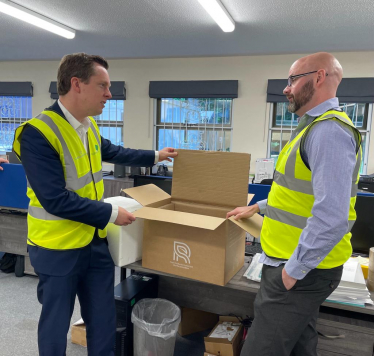 Tom stood talking to an employee of Goggleboxes. They are both wearing hi-viz jackets and are looking at one of the cardboard boxes designed and produced by the business
