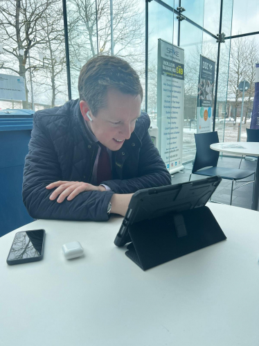 Tom in a virtual meeting with a representative from Vodafone. He is sat down, looking down at his tablet, smiling.