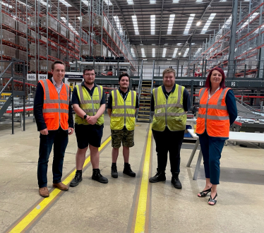 Tom stood with four employees of Wincanton. All are wearing hi-viz jackets and there is extensive warehouse racking in the background.